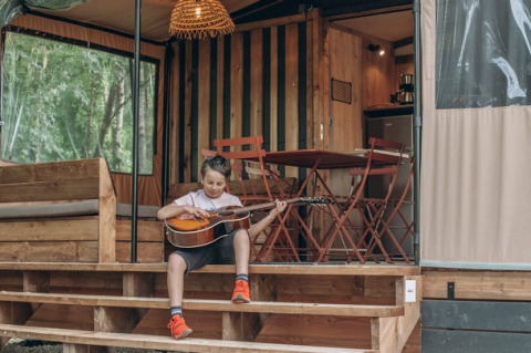 Bambino seduto sui gradini di una tenda safari chiamata Tiny house Galli, suona la chitarra fuori.