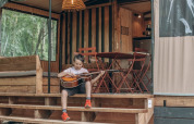Barn sidder på trappen af en safaritelt kaldet Tiny house Galli og spiller guitar foran udendørsområdet.