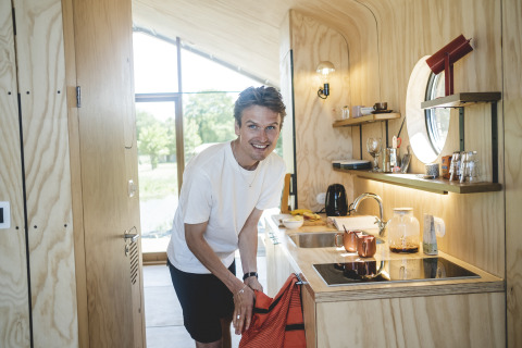 Homme souriant dans une cuisine de lodge moderne en bois clair, vue sur la nature par la fenêtre.
