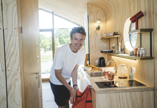 Homme souriant dans une cuisine de lodge moderne en bois clair, vue sur la nature par la fenêtre.