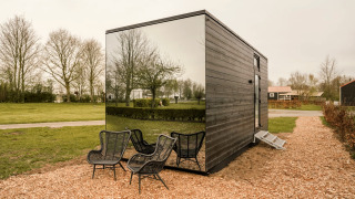 Tiny house with mirrored facade, black outdoor chairs, and natural surroundings with trees