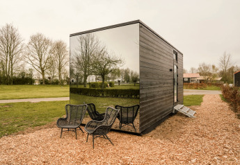 Petite maison avec façade miroir et chaises noires d'extérieur, entourée de verdure et arbres