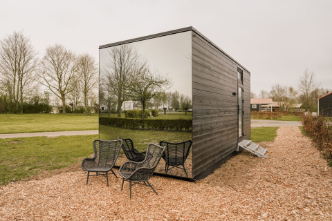 Casa pequeña con fachada espejada y sillas negras al aire libre, rodeada de césped y árboles
