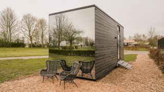 Kleines Haus mit verspiegelter Fassade und schwarzen Stühlen im Freien, umgeben von Natur