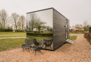 Casa pequeña con fachada espejada y sillas negras al aire libre, rodeada de césped y árboles