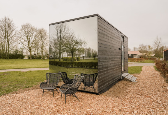 Petite maison avec façade miroir et chaises noires d'extérieur, entourée de verdure et arbres