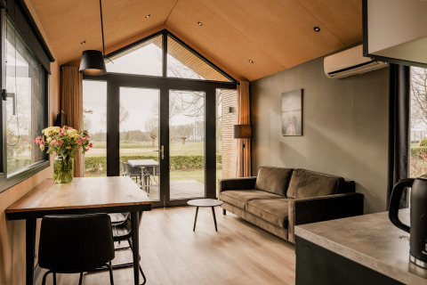 Soggiorno di tiny house moderna con divano, tavolo da pranzo, fiori e vista giardino nei Paesi Bassi.