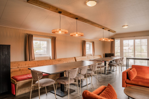 Gezamenlijke lounge met lange tafels, stoelen en oranje banken in Group lodge op Holiday park Eigen Wijze, Nederland.
