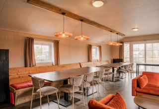 Zona de salón comunal con mesas largas y sofás naranjas en Group lodge de Holiday park Eigen Wijze, Países Bajos.
