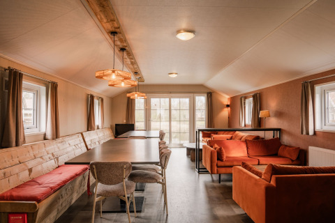 Innenraum der Group Lodge im Holiday Park Eigen Wijze mit langen Tischen, Sofas und Fenstern.