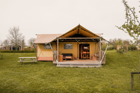 En glamping safari telt med træveranda og picnicbord på en grøn græsplæne, omgivet af natur.
