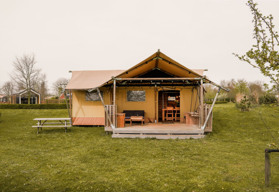 Tenda glamping safari con veranda in legno e tavolo da picnic su prato verde, immersa nella natura.