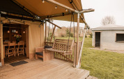 Safari-Glamping-Zelt mit Holzterrasse, Schaukelbank und Blick auf eine runde Hütte im Holiday Park Eigen Wijze.