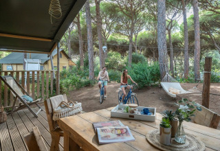 Due persone in bicicletta tra i pini al Sandaya Etruria, parco vacanze in Toscana, Italia, vicino a una veranda.