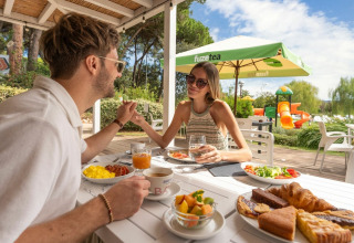 Par nyder morgenmad udendørs på Sandaya Etruria feriepark i Toscana, Italien, med legeplads i baggrunden.