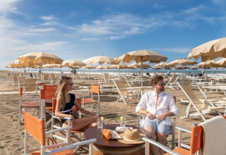 Due persone si rilassano sulla spiaggia di Sandaya Etruria in Toscana tra ombrelloni beige e sedie vuote.
