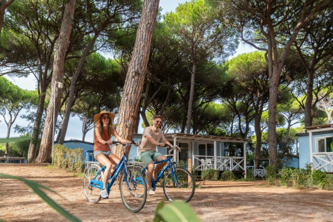 Deux personnes font du vélo dans un parc de vacances avec cabanes en bois et pins, en Toscane, Italie.