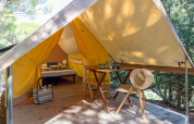 Tienda safari en Sandaya Etruria, Italia, con cama, mesa, sillas y sombrero de paja en terraza de madera.
