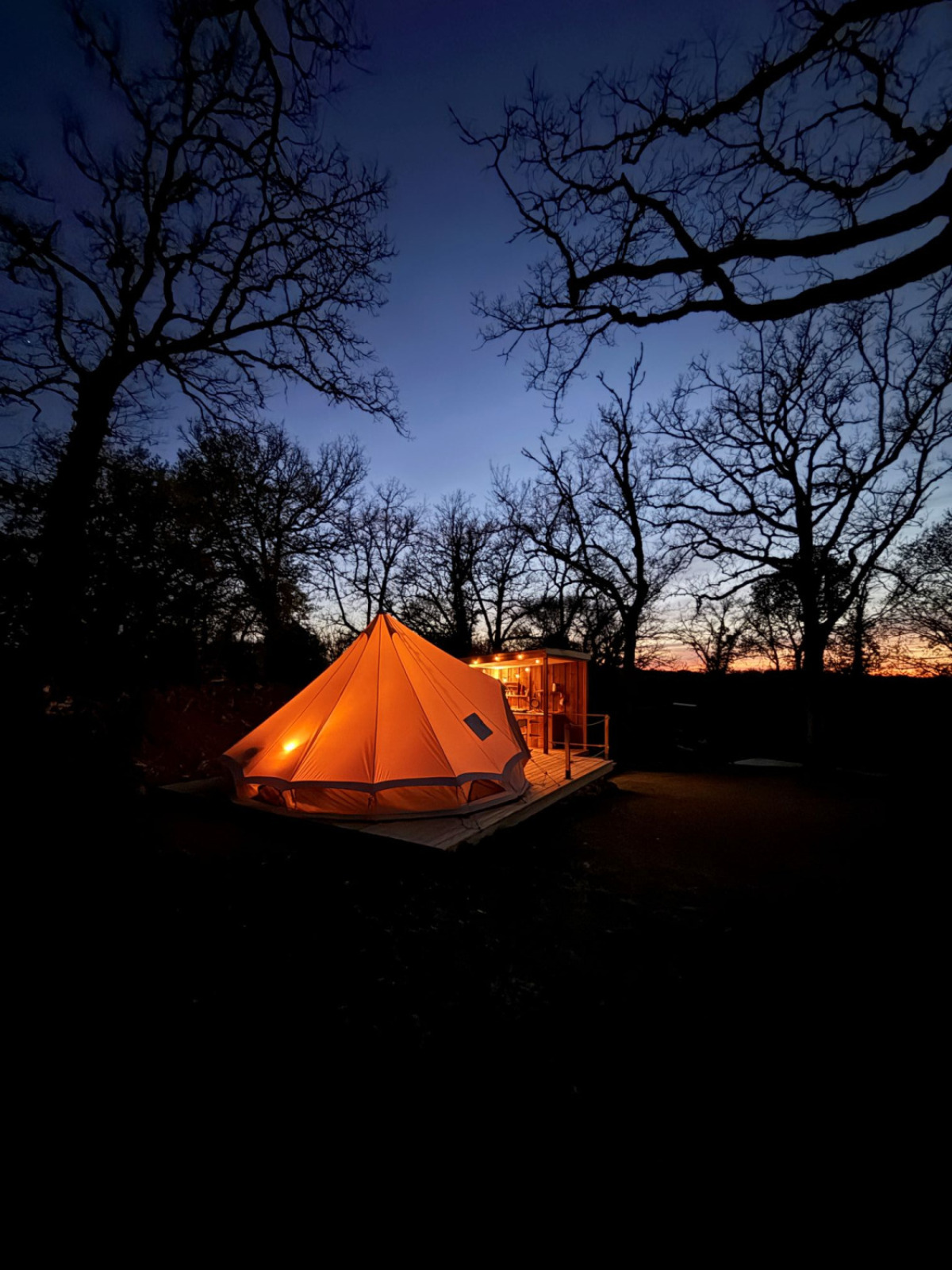 De verlichte Tent Nomad tipi tent bij Camping Domaine de la Faurie in Frankrijk, omgeven door bomen in de avond.