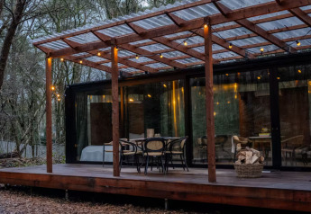 Udendørs terrasse ved Slowlodge Echo lodgen med træmøbler, lyskæder og store glasvinduer i skovmiljø.