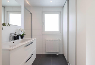Modernes Badezimmer mit weißer Einrichtung, Spiegel, Fenster und Pflanze in der Villa Waterzicht Comfort.