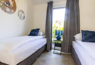 Moderne lodge med to enkeltsenge, blå puder, vindue, gardiner og vægkunst i Holiday park Molenvelden.