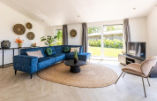 Modernes Wohnzimmer mit blauer Eckcouch, runder Jutematte und großen Fenstern in Villa Waterzicht Luxe.