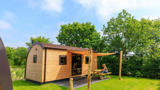 Luxe Woodlodge 4 met houten buitenkant, grasveld, picknicktafel en kind, omringd door groene bomen.
