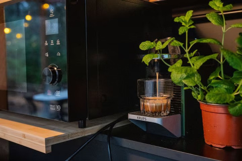 A glass of coffee being brewed on a Krups espresso machine next to a microwave and a mint plant on a shelf.