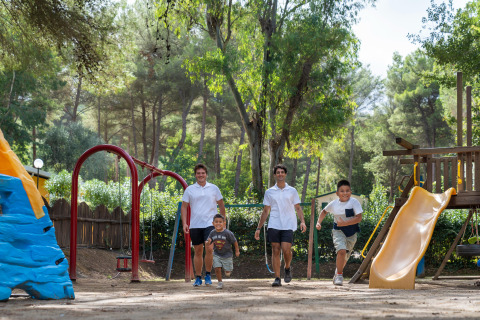 Børn og voksne leger sammen på en legeplads omgivet af træer i Sandaya Le Pianacce feriepark, Toscana.