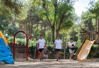 Børn og voksne leger sammen på en legeplads omgivet af træer i Sandaya Le Pianacce feriepark, Toscana.
