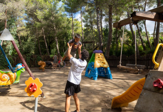Mand løfter barn i legeplads i Sandaya Le Pianacce, Toscana, Italien, omgivet af legeredskaber og træer.