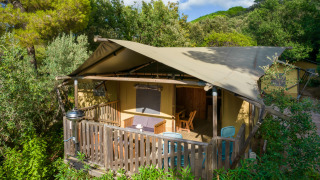 Buitenaanzicht van een Lodge Safari tent met houten veranda, stoelen en omringend groen landschap.