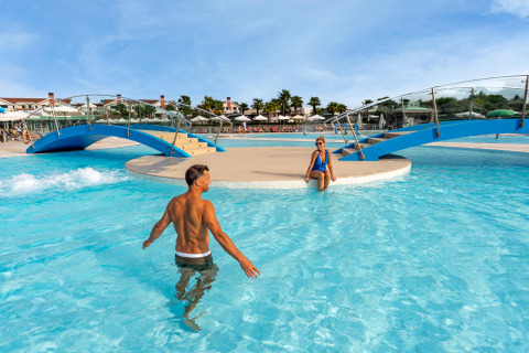 To personer nyder swimmingpoolen ved Sandaya Europa Village feriepark i Veneto, Italien, under en blå himmel.