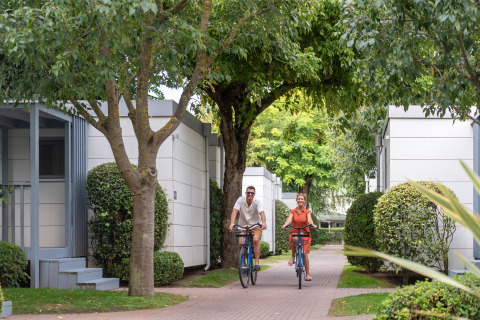 Due persone vanno in bicicletta tra bungalow moderni e alberi nel villaggio turistico Sandaya Europa Village, Veneto.