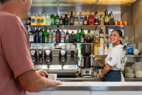 Bartender smiler til kunde i baren på Sandaya Europa Village feriepark i Veneto, Italien, med flasker bag sig.