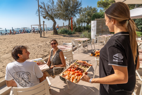 Waitress serving food to a smiling couple at a beachside restaurant in Sandaya Europa Village, Veneto, Italy.