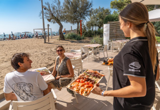 Servering af mad til et smilende par på en udendørs restaurant ved stranden i Sandaya Europa Village, Veneto.