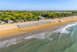 Luftfoto af Sandaya Europa Village ferieparkens strand i Veneto, Italien, med parasoller og grøn skov.