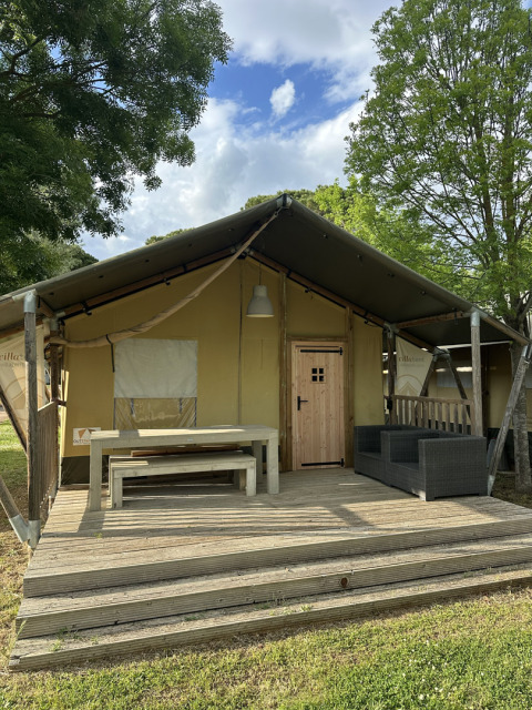 Safaritent Villatent Nomad met houten terras, tafel, banken en sofa, omgeven door bomen en blauwe lucht.