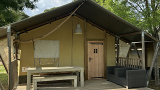Safari tent Villatent Nomad with a wooden deck, table, benches, and sofa, surrounded by trees outdoors.
