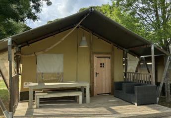 Tienda safari Villatent Nomad con terraza de madera, mesa, bancos y sofá, rodeada de árboles y cielo azul.