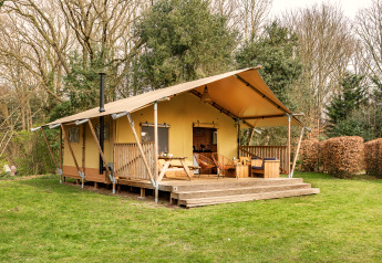 Safari-Zelt mit überdachter Terrasse und Sitzgelegenheiten, umgeben von Bäumen und grünem Rasen.