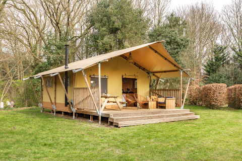Tente safari avec terrasse couverte, mobilier en bois et coin salon extérieur, entourée d’arbres verdoyants.