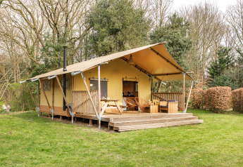 Safari-Zelt mit überdachter Terrasse und Sitzgelegenheiten, umgeben von Bäumen und grünem Rasen.