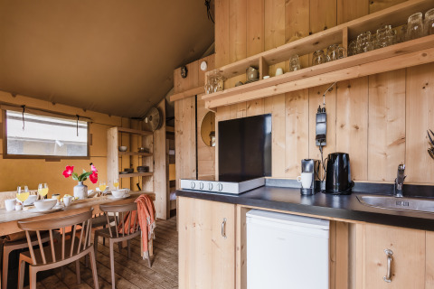 Cuisine moderne et coin repas dans une tente safari, avec mobilier en bois, vaisselle et ustensiles.