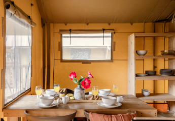 Zona pranzo all’interno di una tenda safari con tavolo apparecchiato, fiori e scaffali con stoviglie.