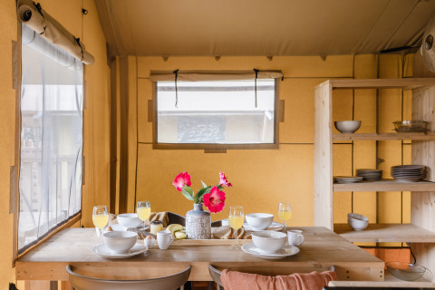 Coin repas dans une tente safari avec table en bois dressée pour le petit-déjeuner, fleurs et vaisselle.