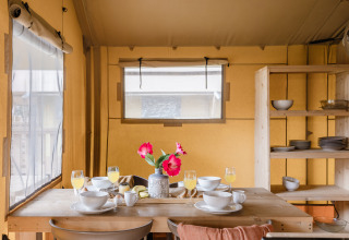 Coin repas dans une tente safari avec table en bois dressée pour le petit-déjeuner, fleurs et vaisselle.