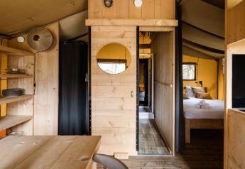 Innenraum eines Safarizeltes mit Holzmöbeln, Schlafzimmer, offenen Regalen und gemütlicher Atmosphäre.